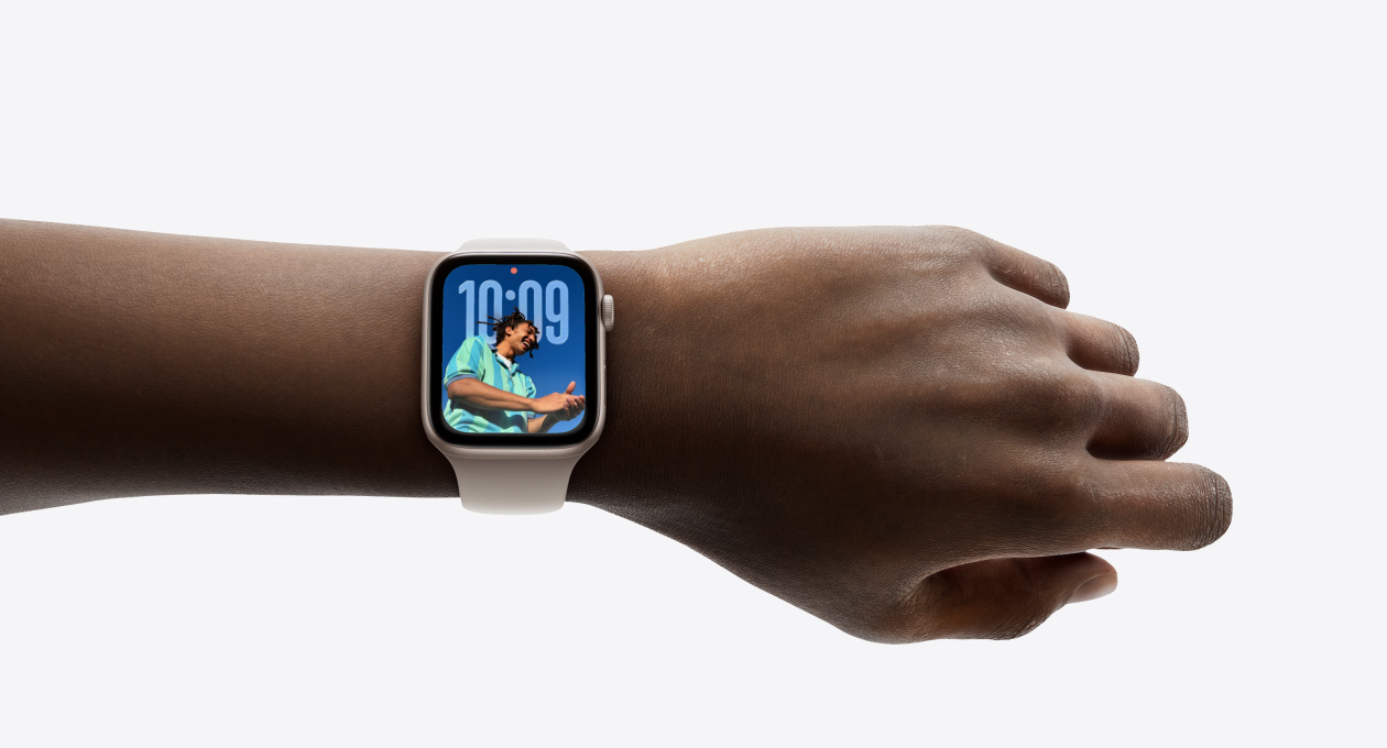 Foto close-up Apple Watch berwarna Perak dengan tali jam karet di pergelangan tangan seseorang, menampilkan aplikasi Waktu dalam desain Liquid Glass baru