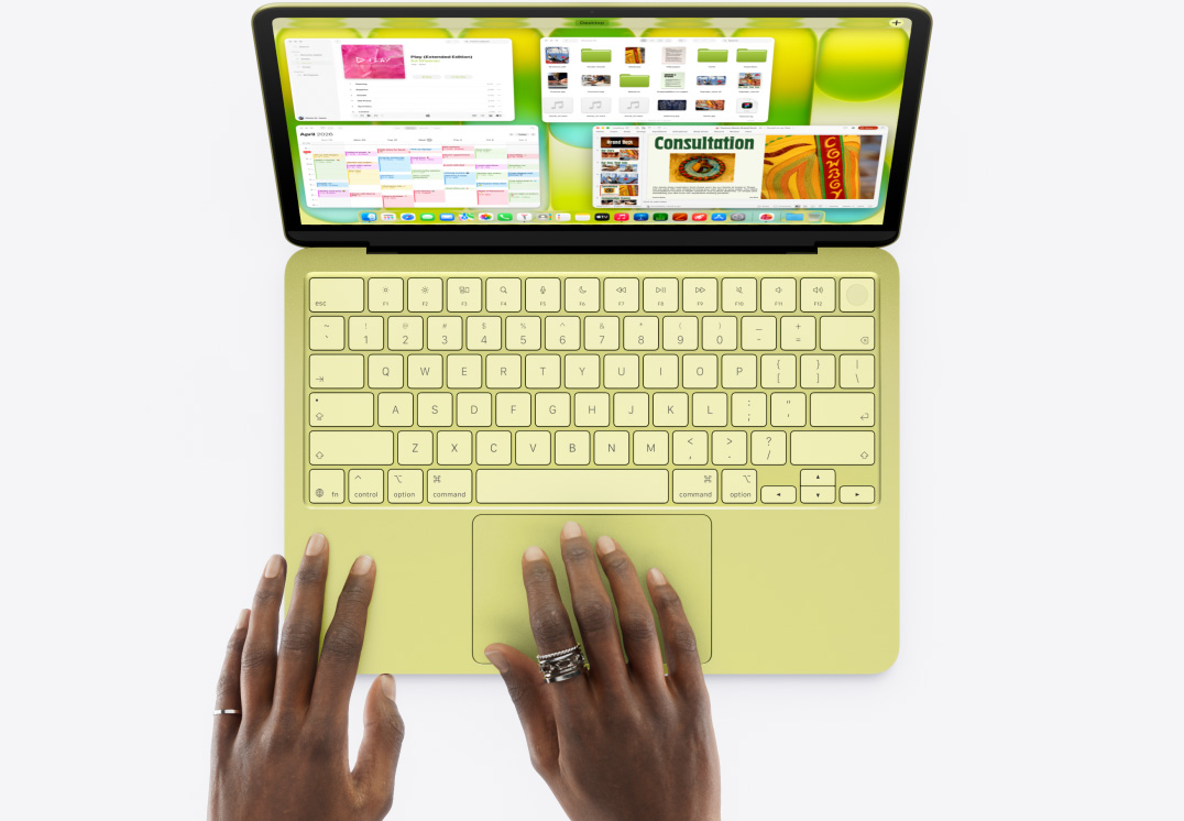 Citrusų spalvos „MacBook Neo“, manipuliatorių sukiojantys pirštai, atidaryti keli langai, įskaitant „Music“, „Kalendorius“ ir „Microsoft PowerPoint“ programas