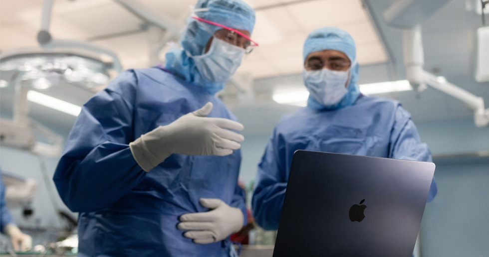 Kaksi kirurgia leikkaussalissa katsoo MacBookia. Toinen elehtii käsillään ja toinen tarkkailee