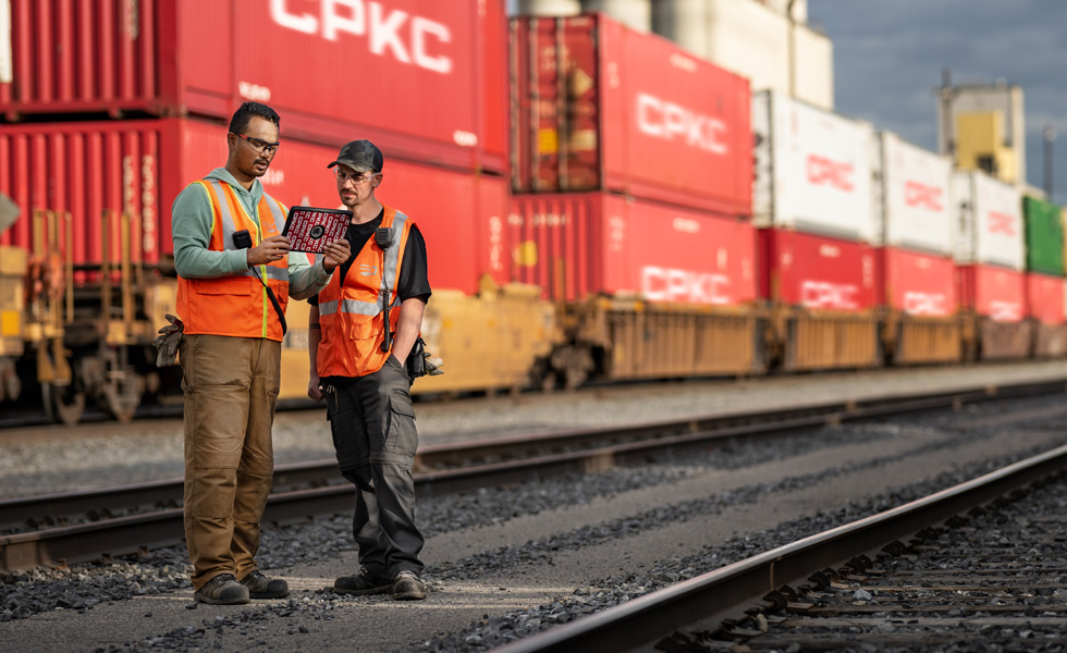 Two railway workers reference an iPad while standing in a train depot beside a stationary train carrying shipping containers