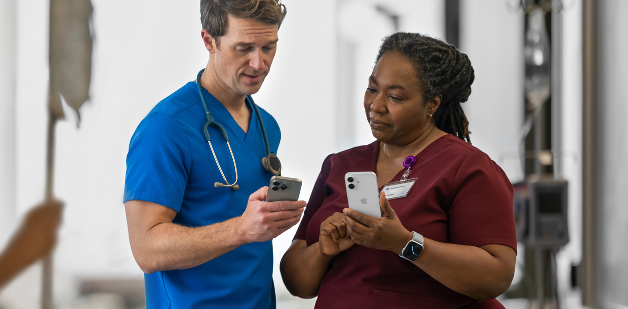 En mannlig behandler i blå uniform ser på en iPhone og viser noe på den til en kvinnelig behandler i rød uniform, som også har en iPhone.