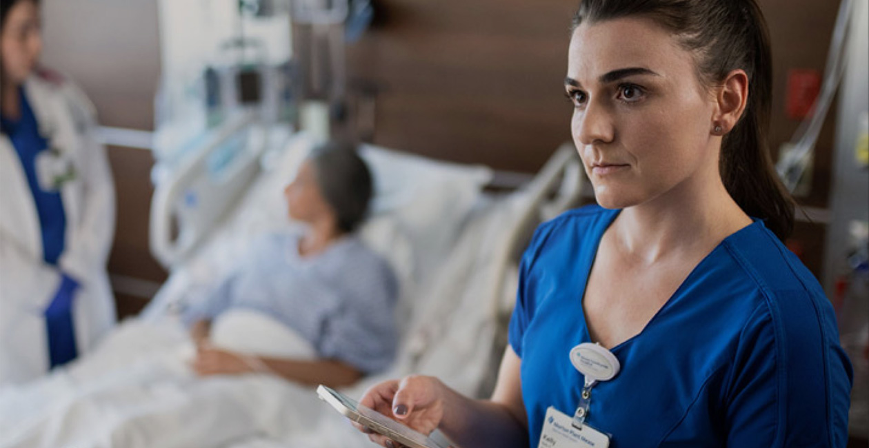 Une infirmière envoie un message sur son iPhone pendant qu’un autre prestataire de soins s’entretient avec une personne hospitalisée.